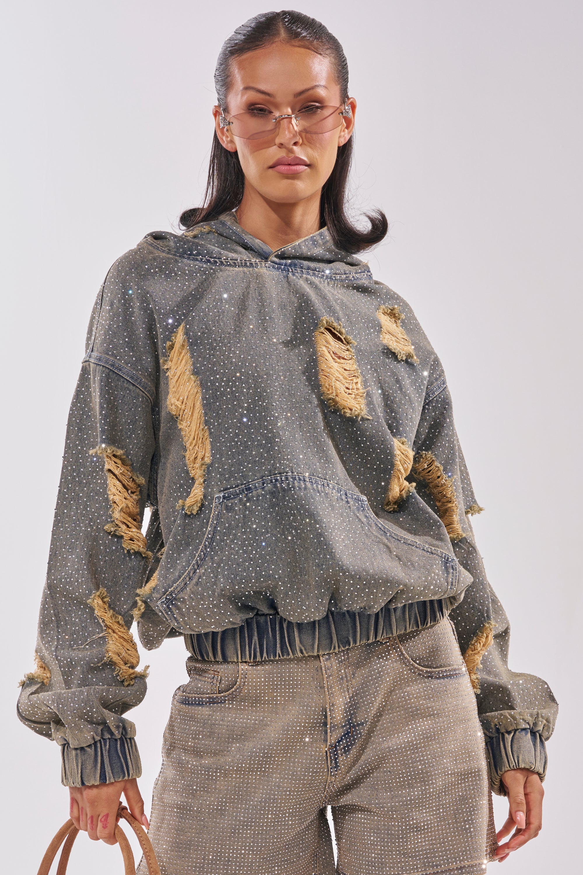 Wearing the DEMI RHINESTONE DENIM HOODIE with distressed gray fabric and matching pants, a woman in clear-rimmed glasses poses with a tan handbag against a light background. 3