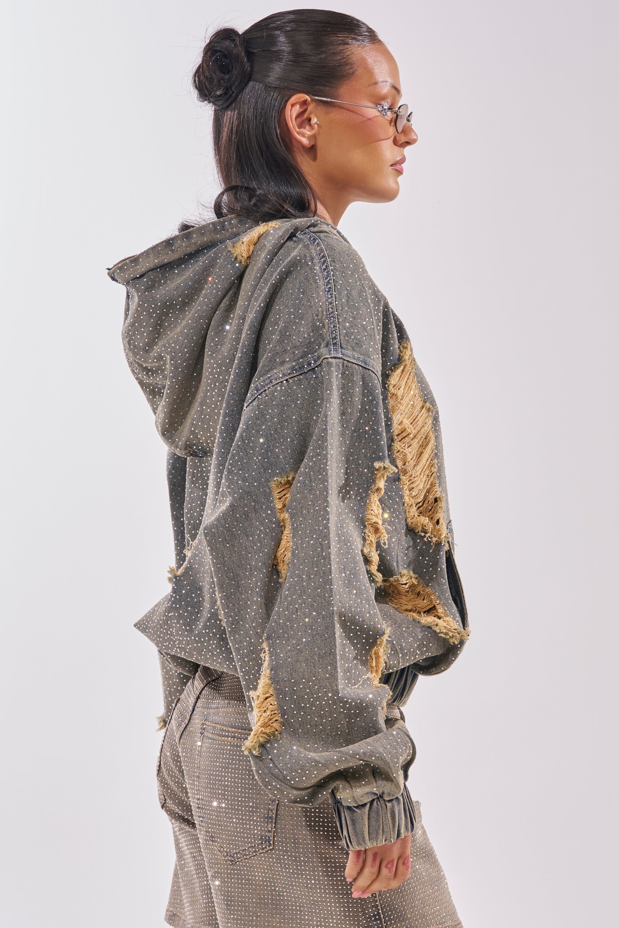 Wearing the DEMI RHINESTONE DENIM HOODIE with frayed patches and glasses, a person with dark hair in a bun pairs it with studded pants, creating a striking festival streetwear look against a light background. 4