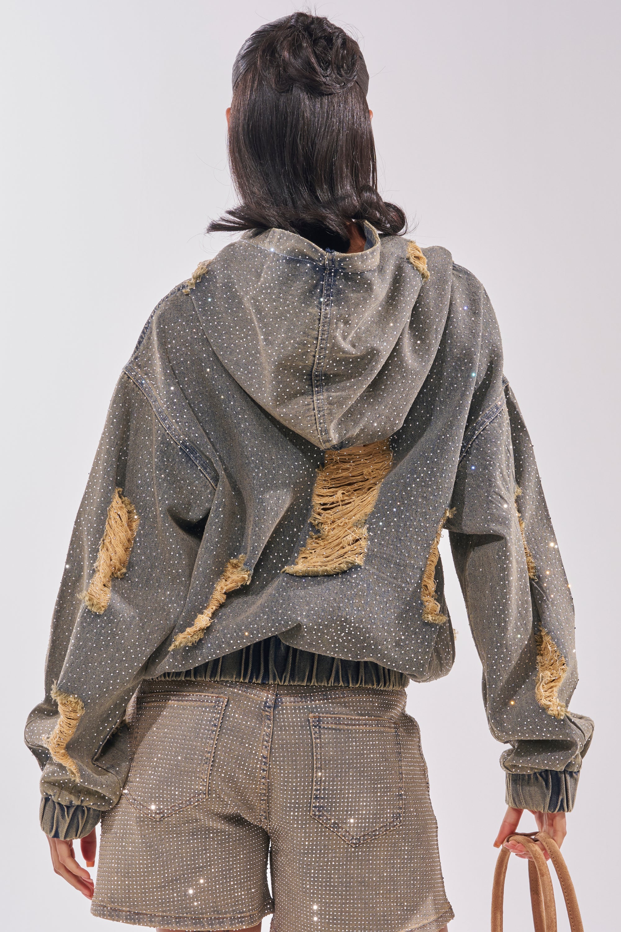 From behind, a woman sports the DEMI RHINESTONE DENIM HOODIE in greenish distressed denim with sparkling rhinestones, matching shorts, and tan bag—an edgy festival streetwear look featuring large frayed rips. 5