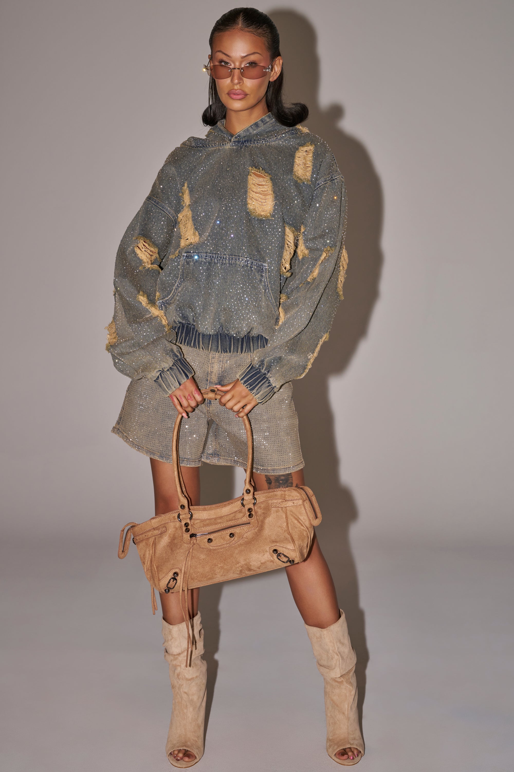 A woman poses in festival streetwear, wearing the DEMI RHINESTONE DENIM HOODIE with matching shorts, a tan suede handbag, tan open-toe boots, and sunglasses, standing against a plain backdrop. 7