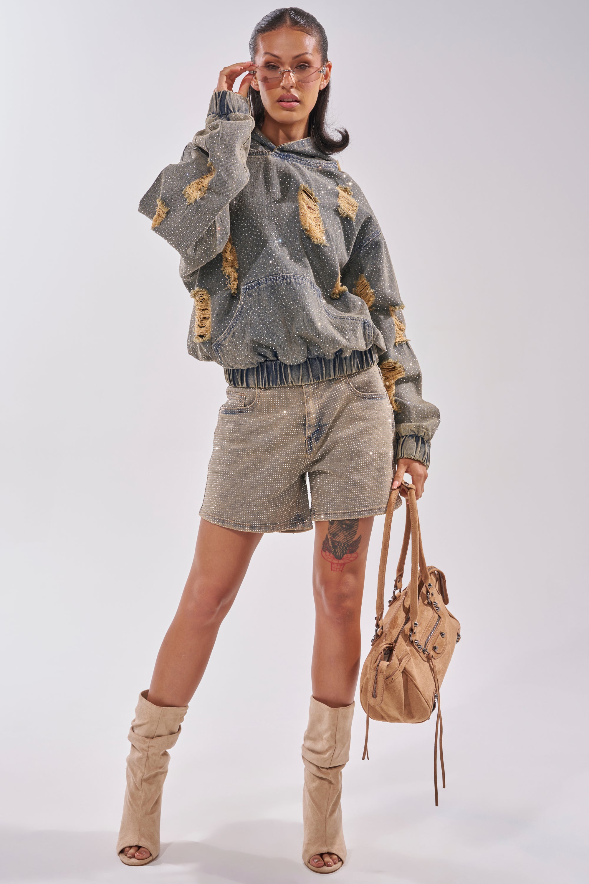 A woman poses in the DEMI RHINESTONE DENIM HOODIE and matching shorts, paired with tan slouchy boots and a tan handbag. She wears sunglasses, hair styled back, showcasing festival streetwear against a plain light background. 8