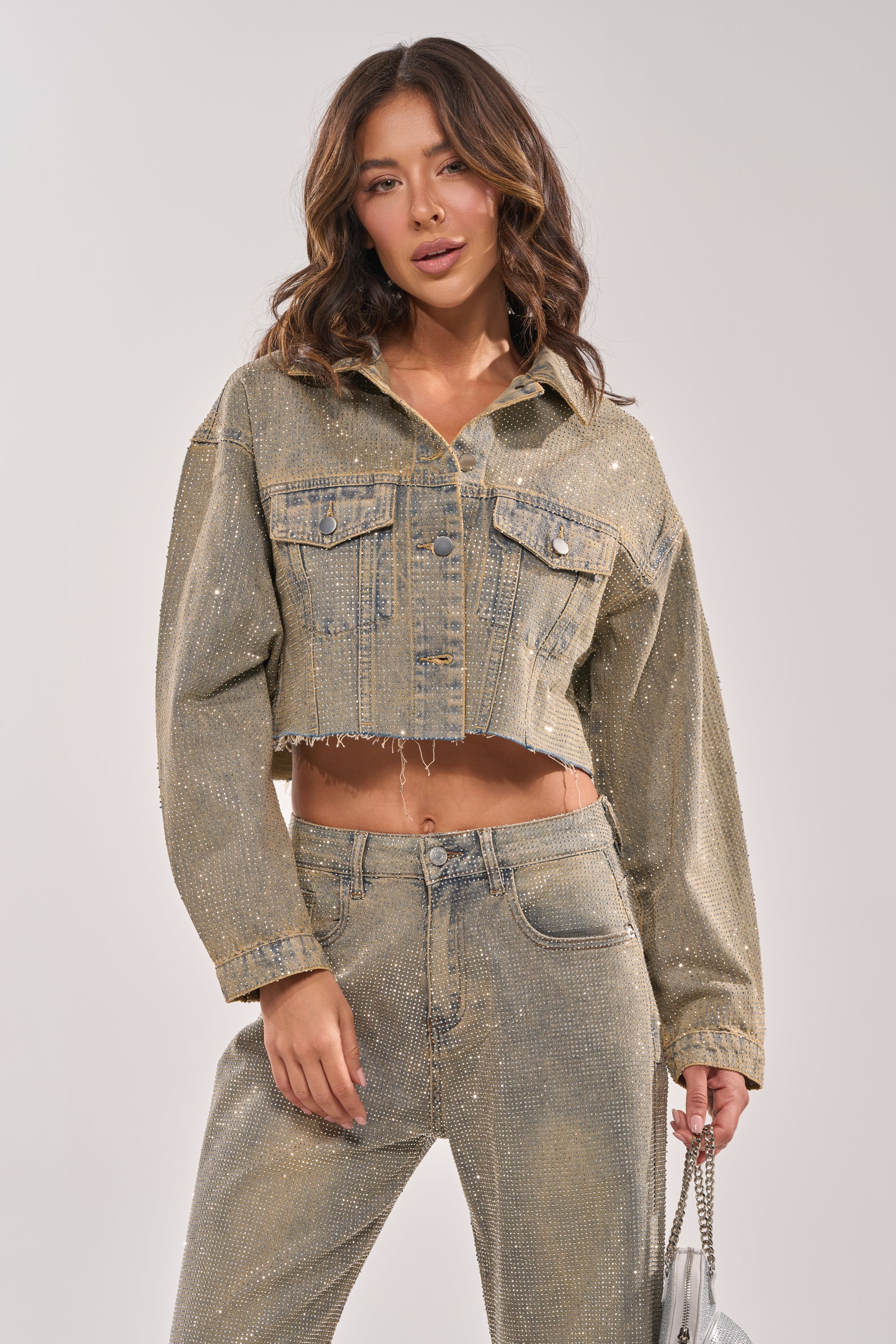 A woman with wavy brown hair wears the DEMI EMBELLISHED DENIM JACKET and matching high-waisted jeans, both adorned with subtle rhinestones. She holds a small silver handbag and poses against a plain light background. 5