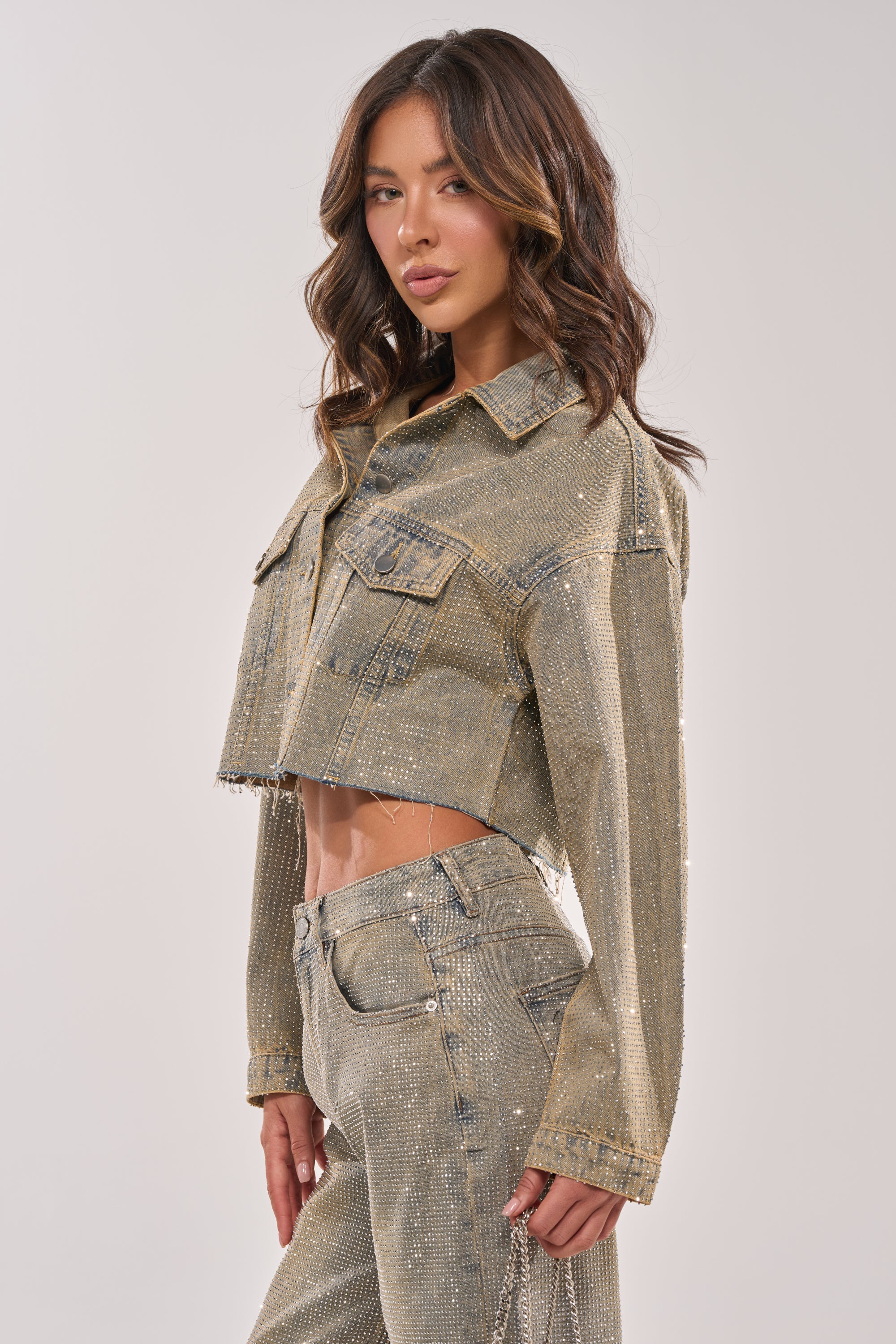 A woman with wavy brown hair confidently poses against a plain light background, wearing the DEMI EMBELLISHED DENIM JACKET—a sparkling, metallic cropped denim piece paired with matching pants. 7