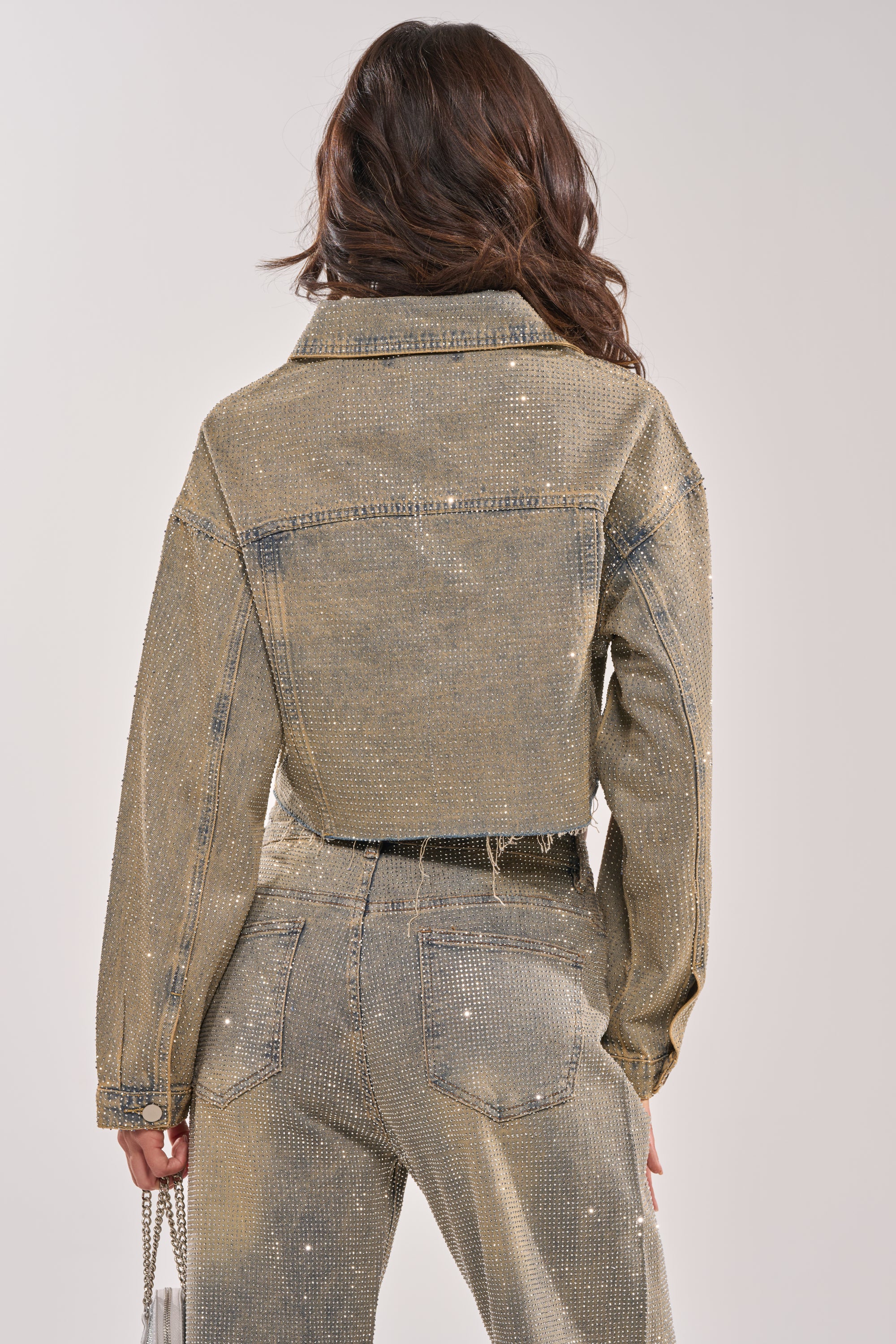 A woman is seen from the back in the DEMI EMBELLISHED DENIM JACKET, paired with high-waisted sparkling pants. She carries a silver chain purse in her left hand. 8