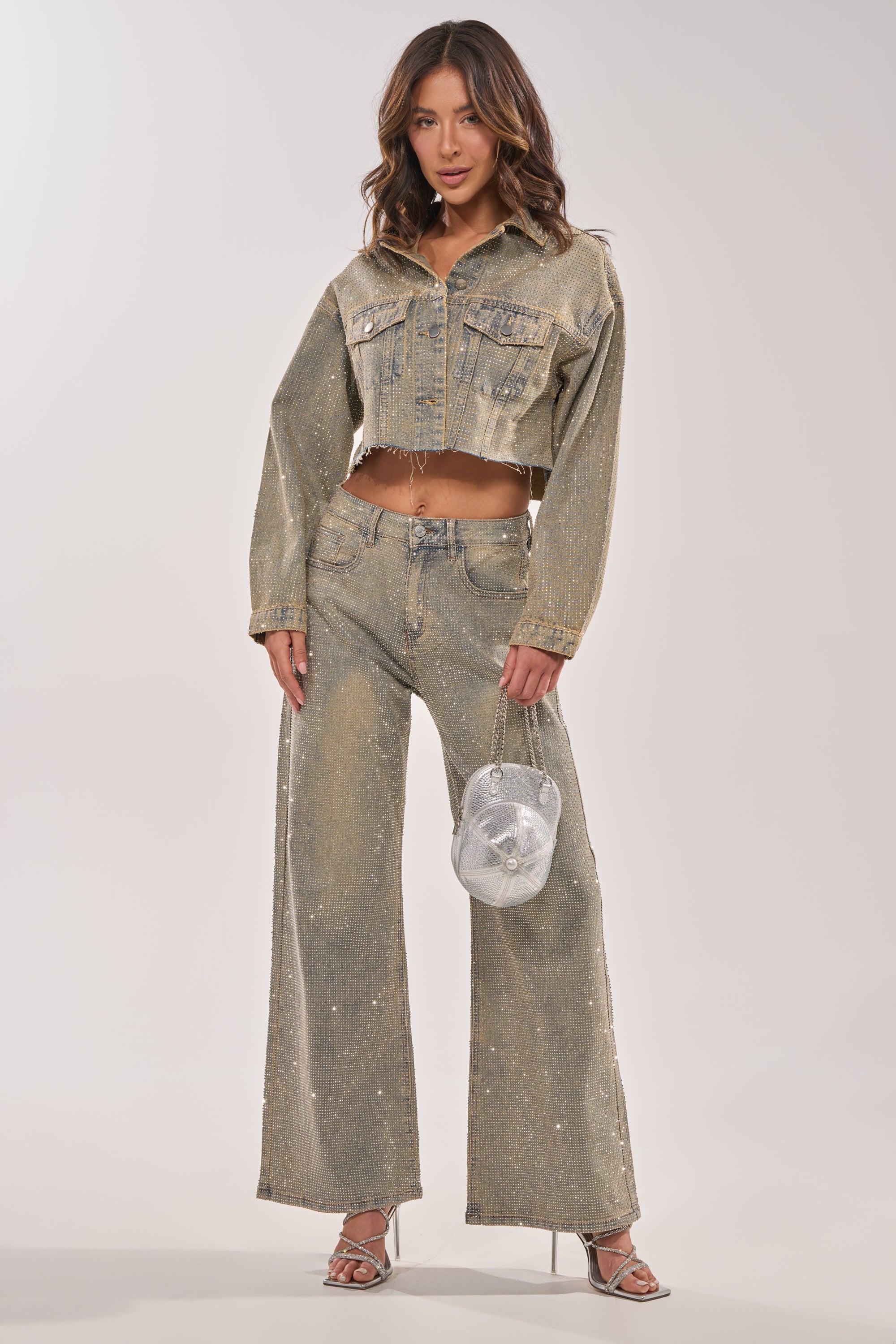 A woman poses confidently in the DEMI EMBELLISHED DENIM JACKET and matching wide-leg jeans, holding a shiny silver bag and wearing silver heels. Her soft waves frame her face as she stands against a plain light background. 9