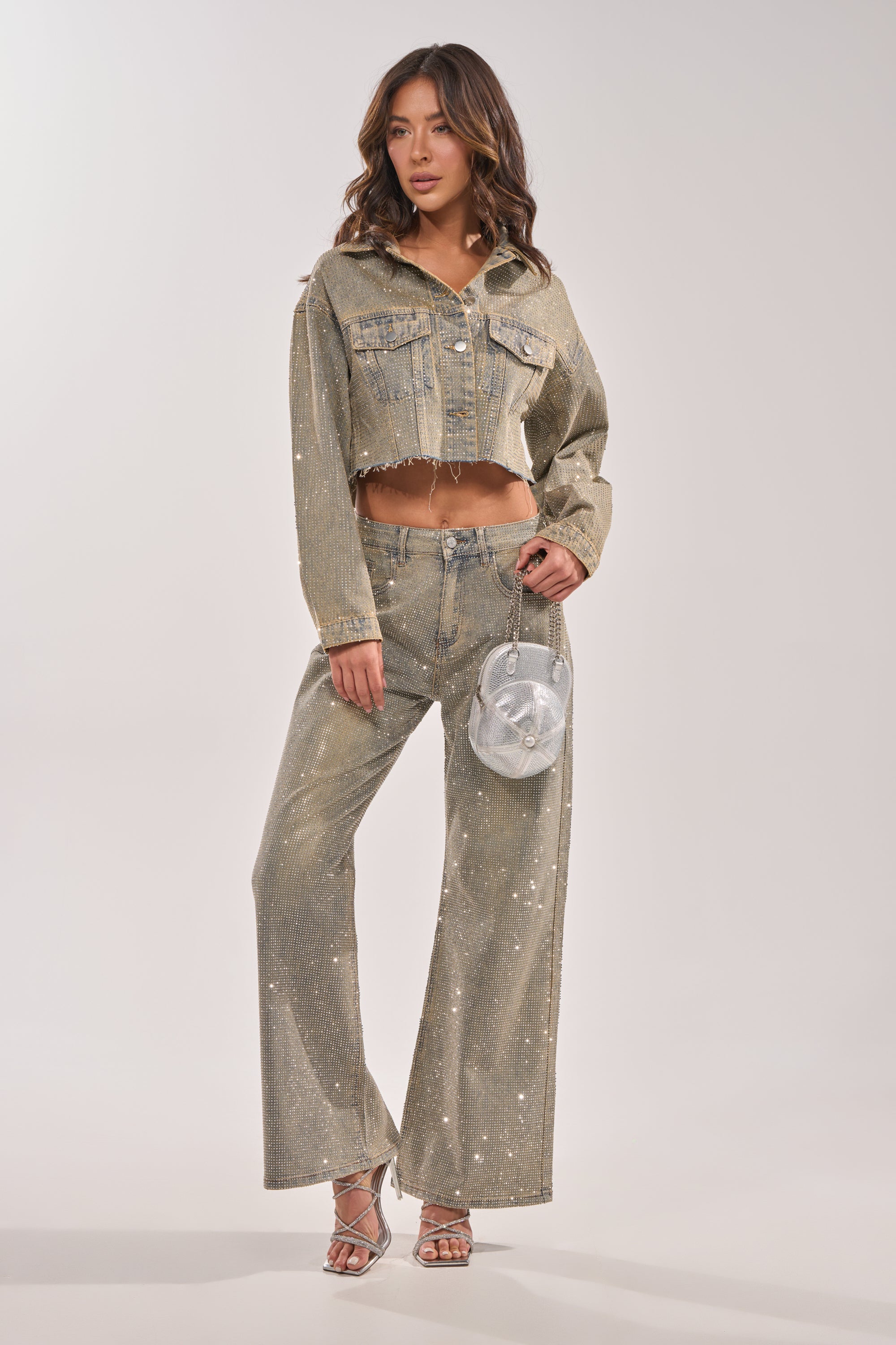 A woman in the DEMI EMBELLISHED DENIM JACKET and wide-leg metallic jeans, holding a small round silver purse and wearing silver strappy heels, stands against a plain light background. 10