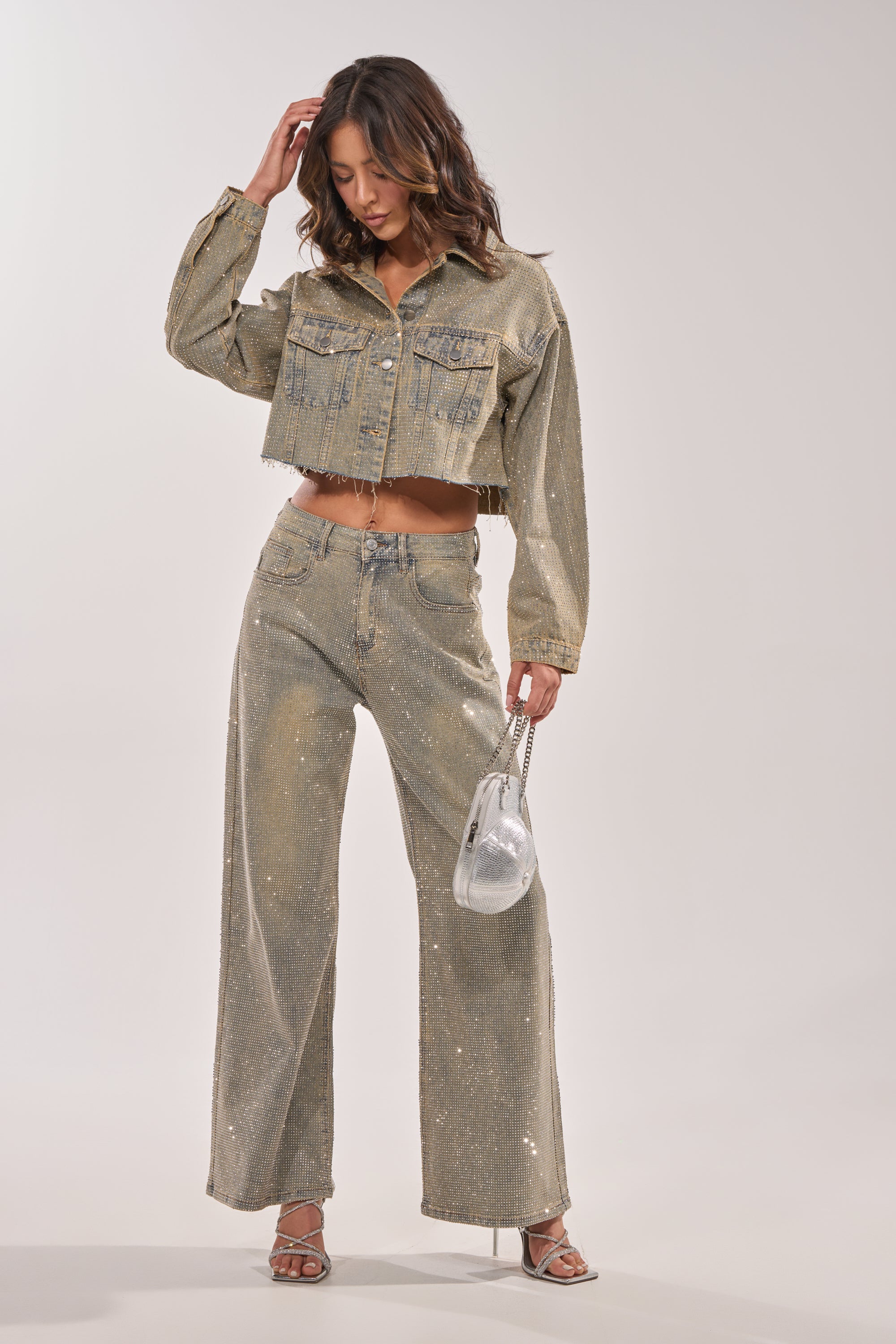 A woman poses in the DEMI EMBELLISHED DENIM JACKET with matching wide-leg jeans, holding a transparent handbag and wearing silver high-heeled sandals. 11