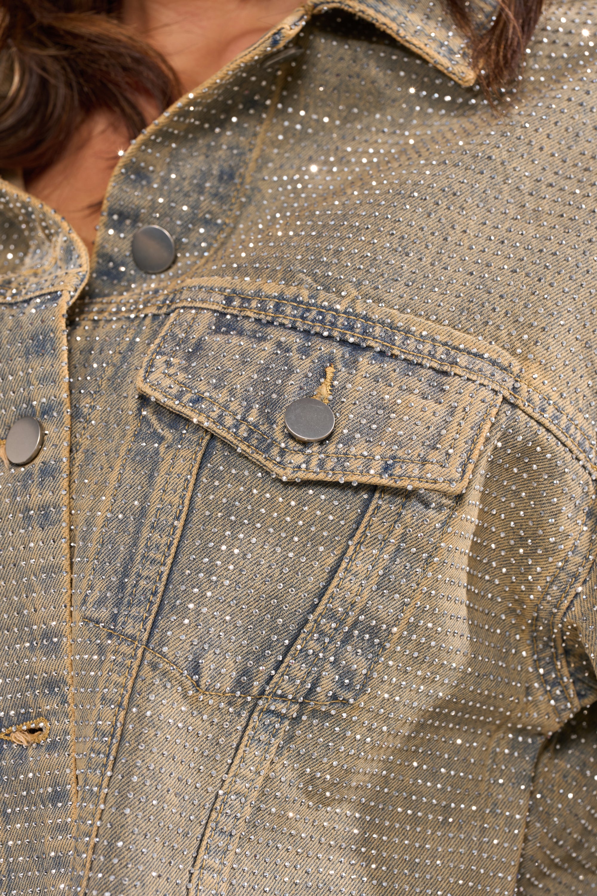 Close-up of the DEMI EMBELLISHED DENIM JACKET featuring silver rhinestones, metal buttons, and a front pocket flap. The cropped jacket is worn by someone with long, wavy hair partially visible. 12