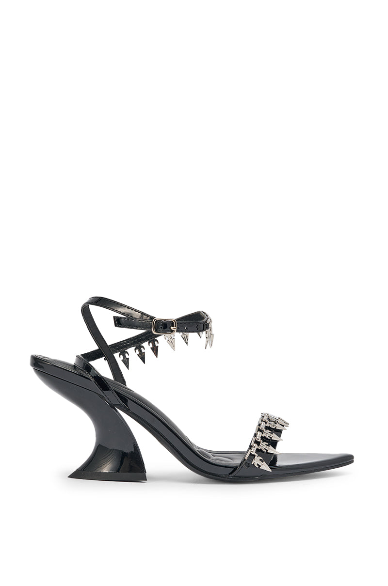 The AKIRA GOLD DEMISE sandal features black faux patent leather, silver hardware embellishments on ankle and toe straps, a chunky flared heel, and an ankle strap, shown against a white background. 6