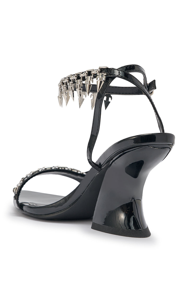The photo shows the AKIRA GOLD DEMISE sandal in black faux patent leather, featuring silver hardware, chunky straps, a curved heel, and ankle strap with metallic spike and chain embellishments, viewed from the back. 4