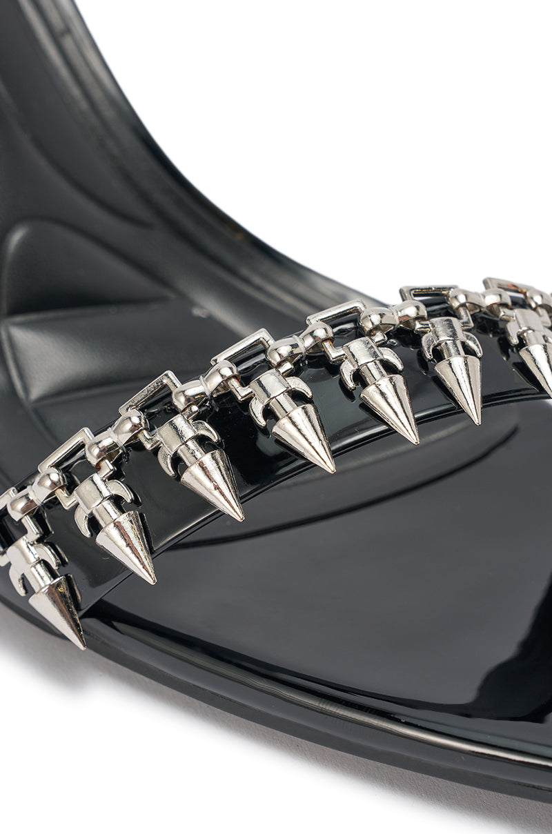 Close-up of the AKIRA GOLD DEMISE sandal in shiny black faux patent leather, featuring chunky strappy design adorned with silver hardware and chain-like embellishments. 7