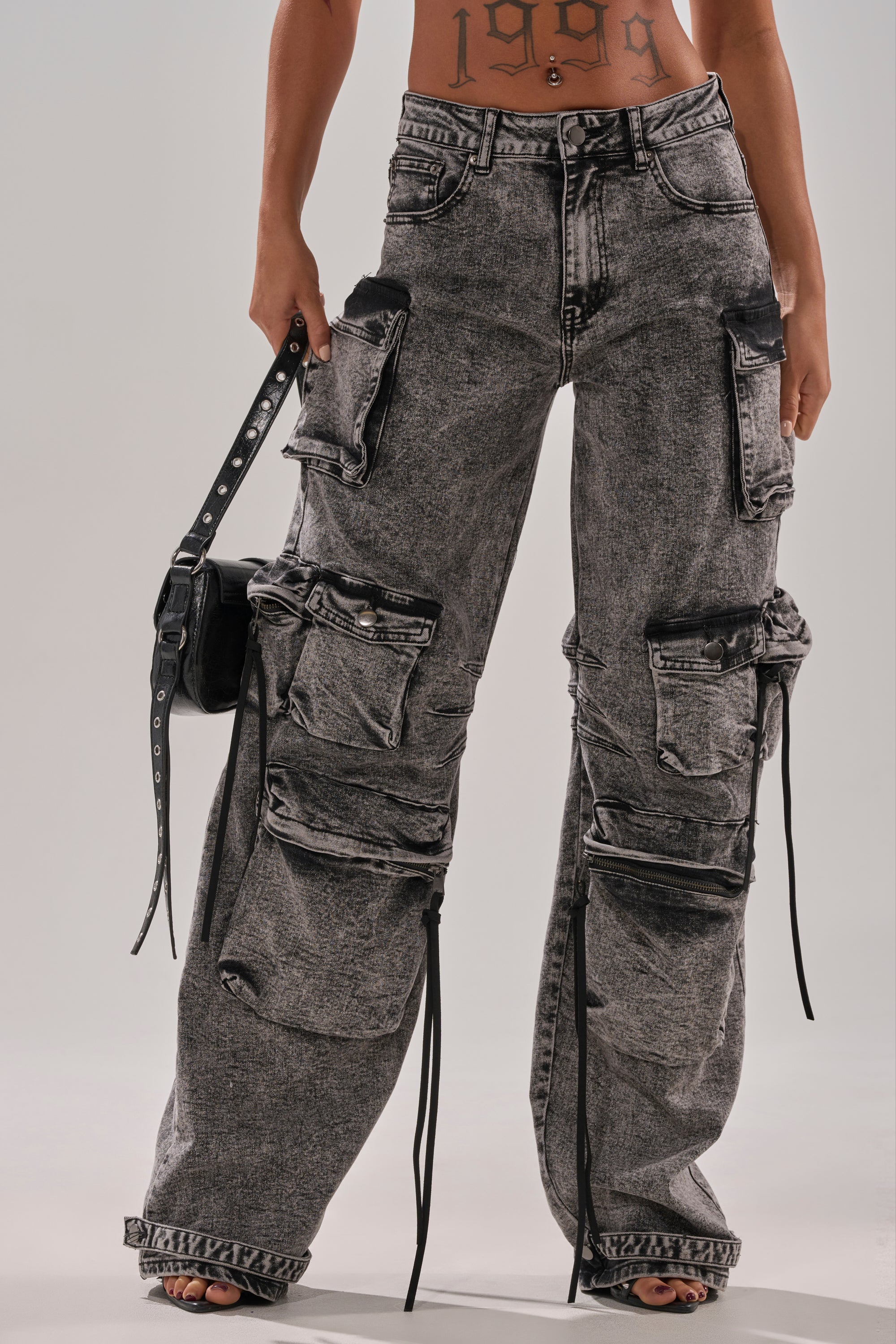 A person wears COOL GIRL DENIM CARGO PANT—wide-leg, acid-washed with black straps and multiple large pockets—holding a black purse. Only their tattooed torso and legs are visible.