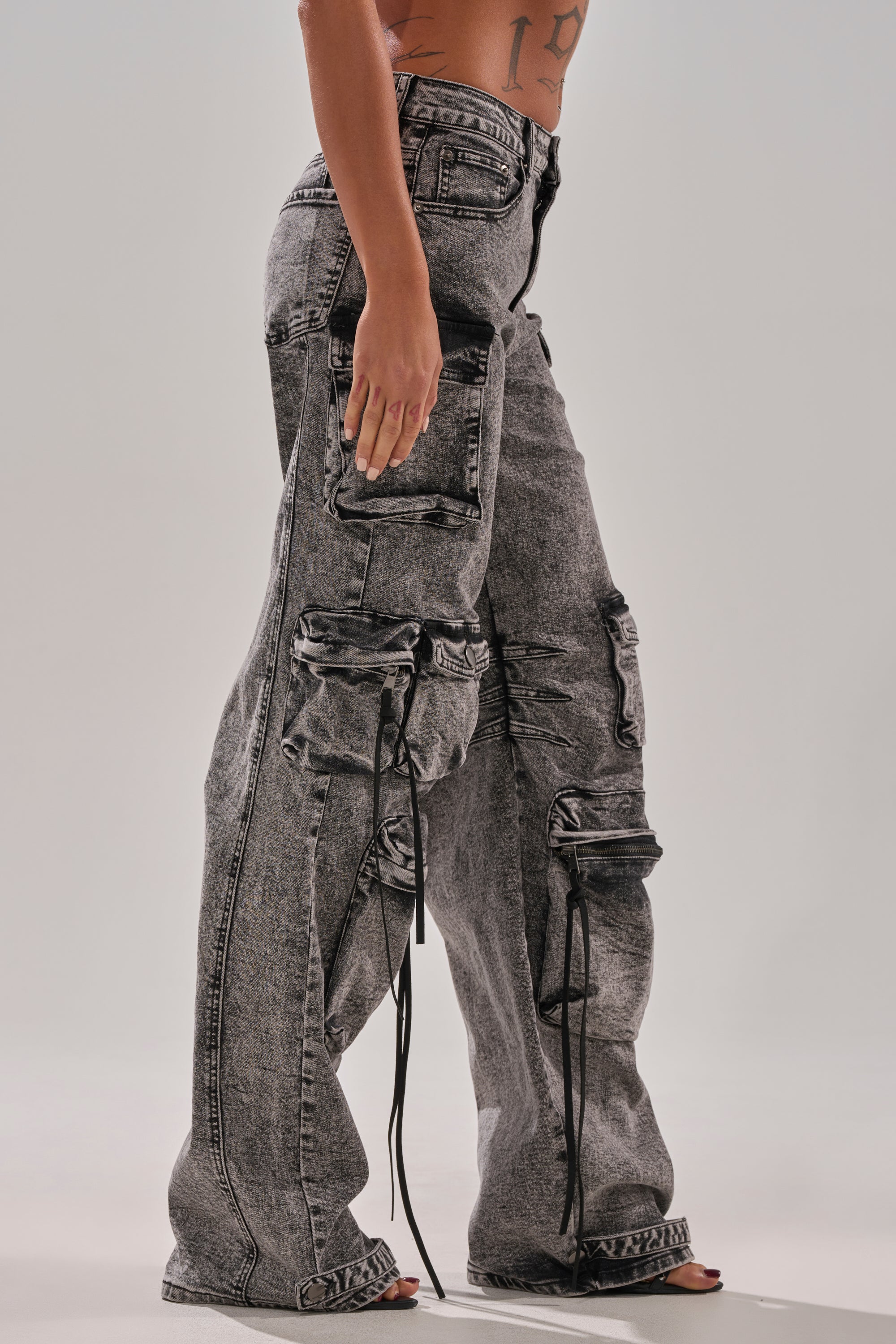 A person models the COOL GIRL DENIM CARGO PANT—high-waisted, wide-leg acid washed jeans featuring multiple pockets, drawstrings, and zippers—while standing barefoot against a plain background.