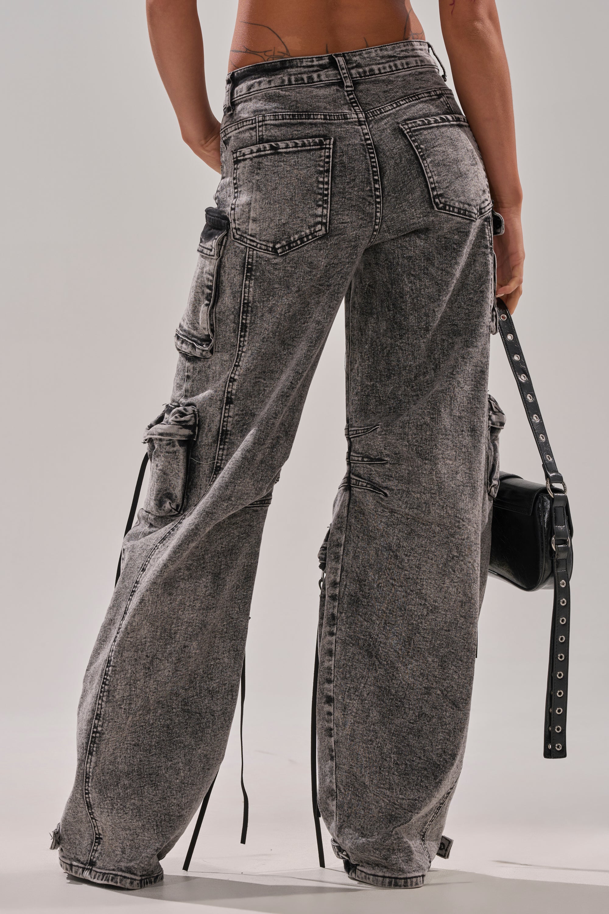 A person stands with their back to the camera, holding a black handbag and wearing COOL GIRL DENIM CARGO PANT—loose-fitting, high-waisted jeans with multiple pockets and a wide-leg, acid-washed silhouette.
