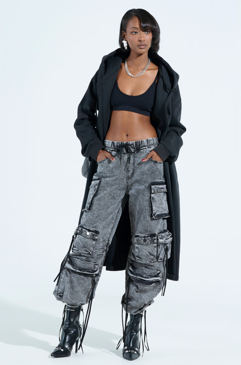A person confidently wears a black crop top, long black coat, and LOOKS FIRE DENIM JOGGER pants with multiple pockets and tassel details. They accessorize with a necklace and black heeled boots against a plain white background.