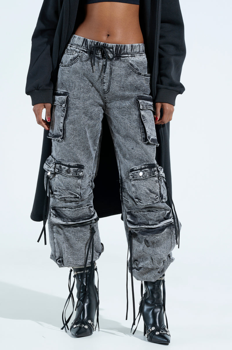 A person in a long black coat and gray LOOKS FIRE DENIM JOGGER pants, featuring multiple cargo pockets and drawstrings. They're also in black high-heeled boots, standing against a plain white background that accentuates the utilitarian flair of their attire.