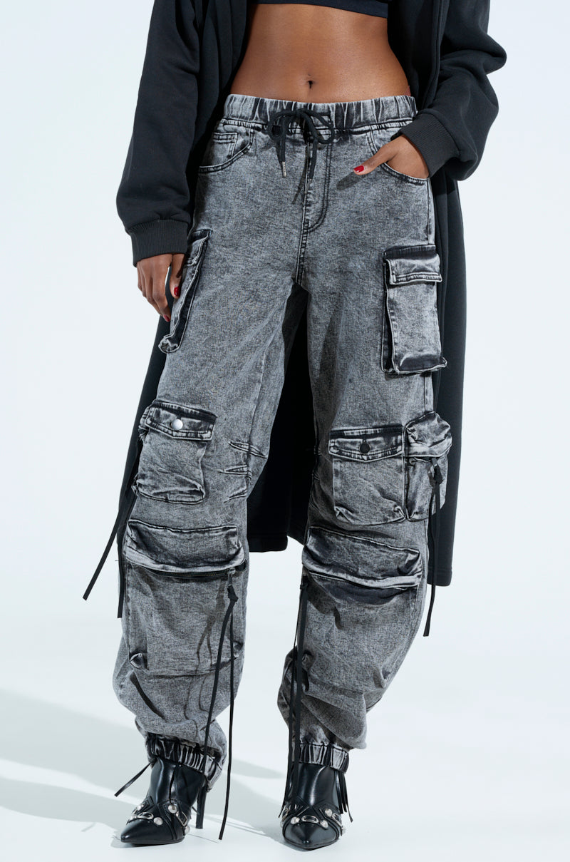 A person in gray cargo pants with multiple pockets and drawstrings, paired with a black long-sleeve top and shoes stands against a plain background, seamlessly blending utility and style like the LOOKS FIRE DENIM JOGGER.