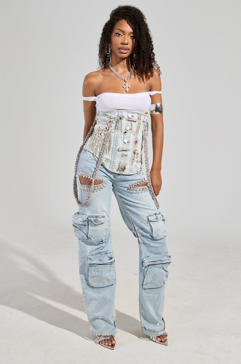 A woman with curly hair stands confidently in a white off-shoulder top and the CALLING THE SHOTS CARGO DENIM WITH RHINESTONES, featuring light blue, rhinestone-embellished cargo denim pants with a high-rise waist. She accessorizes with silver jewelry, including a necklace, bracelets, and a chain. The background is plain gray.