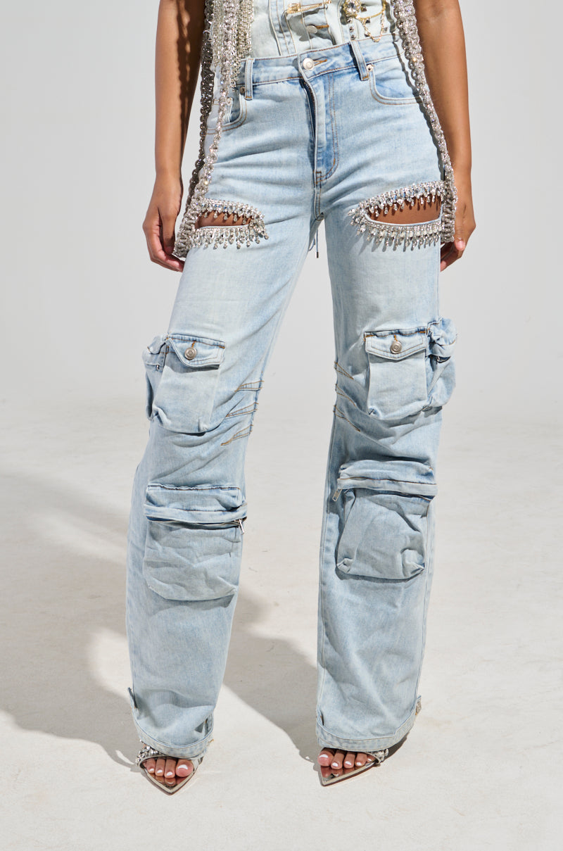 A person wearing the CALLING THE SHOTS CARGO DENIM WITH RHINESTONES in light blue, featuring a high-rise waist, cutouts, decorative chains at the thighs, and multiple cargo-style pockets embellished with subtly glistening rhinestones. The background is a plain light gray.