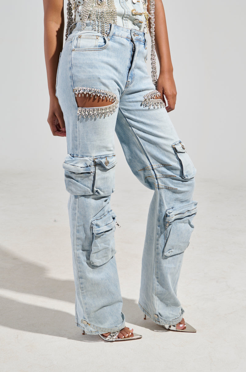 A person wearing the CALLING THE SHOTS CARGO DENIM WITH RHINESTONES, featuring a high-rise waist and multiple pockets. The light blue denim is adorned with dazzling rhinestones and cutouts above the knees with dangling crystals, creating a perfect match for silver high heels.
