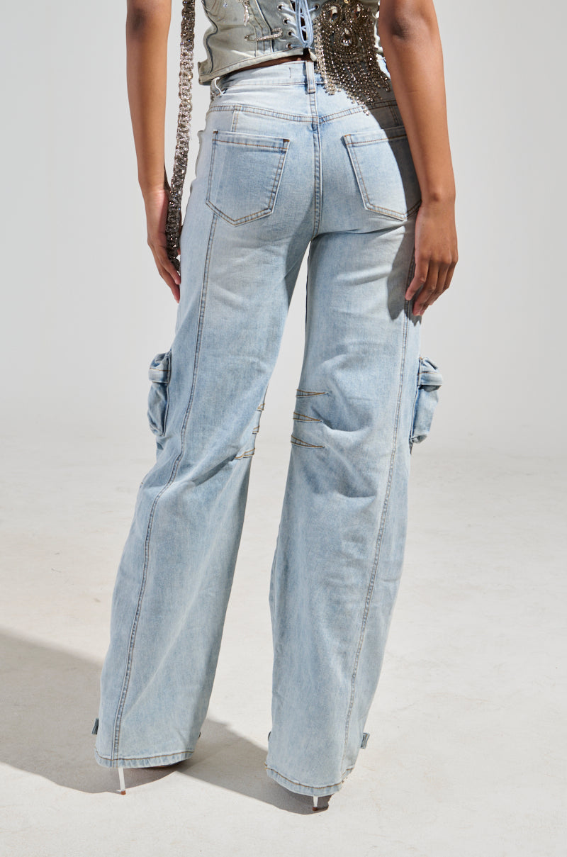 Someone is dressed in the CALLING THE SHOTS CARGO DENIM WITH RHINESTONES, featuring light blue cargo pants with a high-rise waist and prominent rear and side pockets. The partially visible top boasts a rhinestone-embellished texture. The scene is softly lit against a plain background.