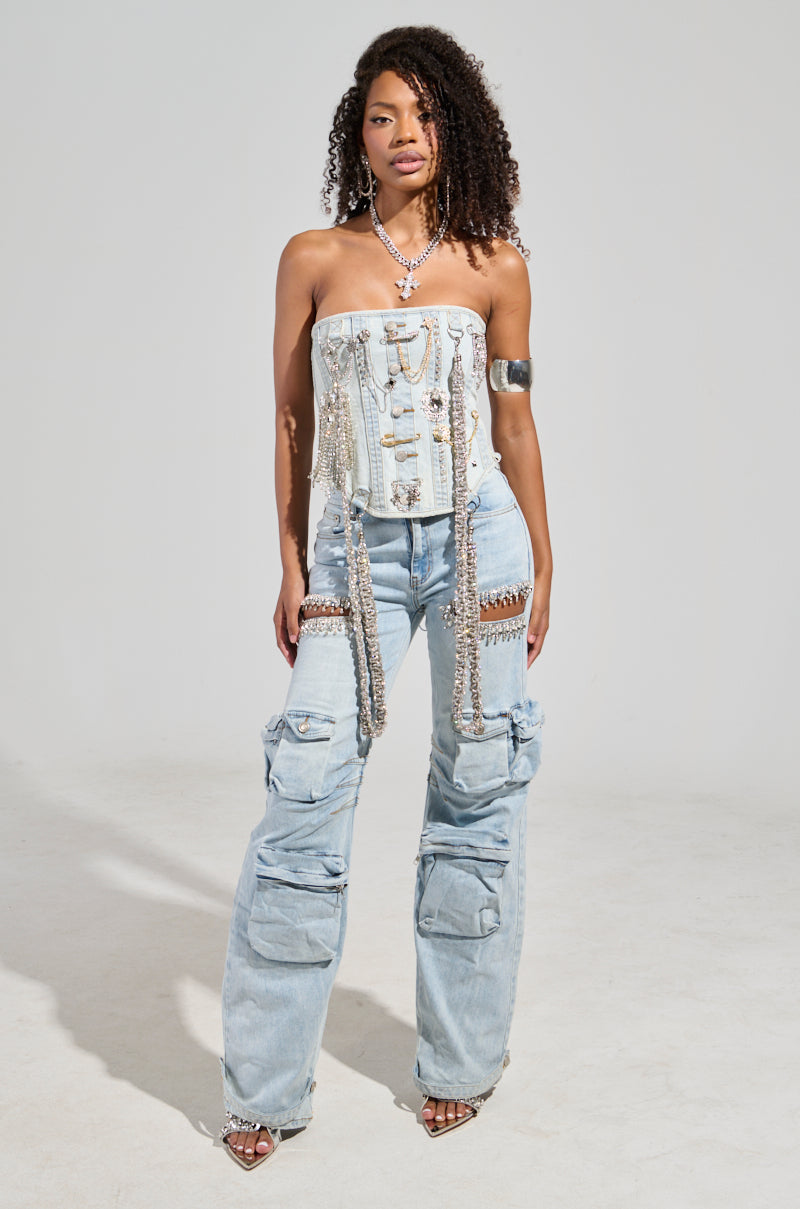 A person with curly hair stands confidently, wearing a strapless denim top adorned with chains and layered necklaces. They pair it with the CALLING THE SHOTS CARGO DENIM WITH RHINESTONES jeans, which have a high-rise waist and large pockets. An arm cuff graces their left arm as they pose against a plain background.