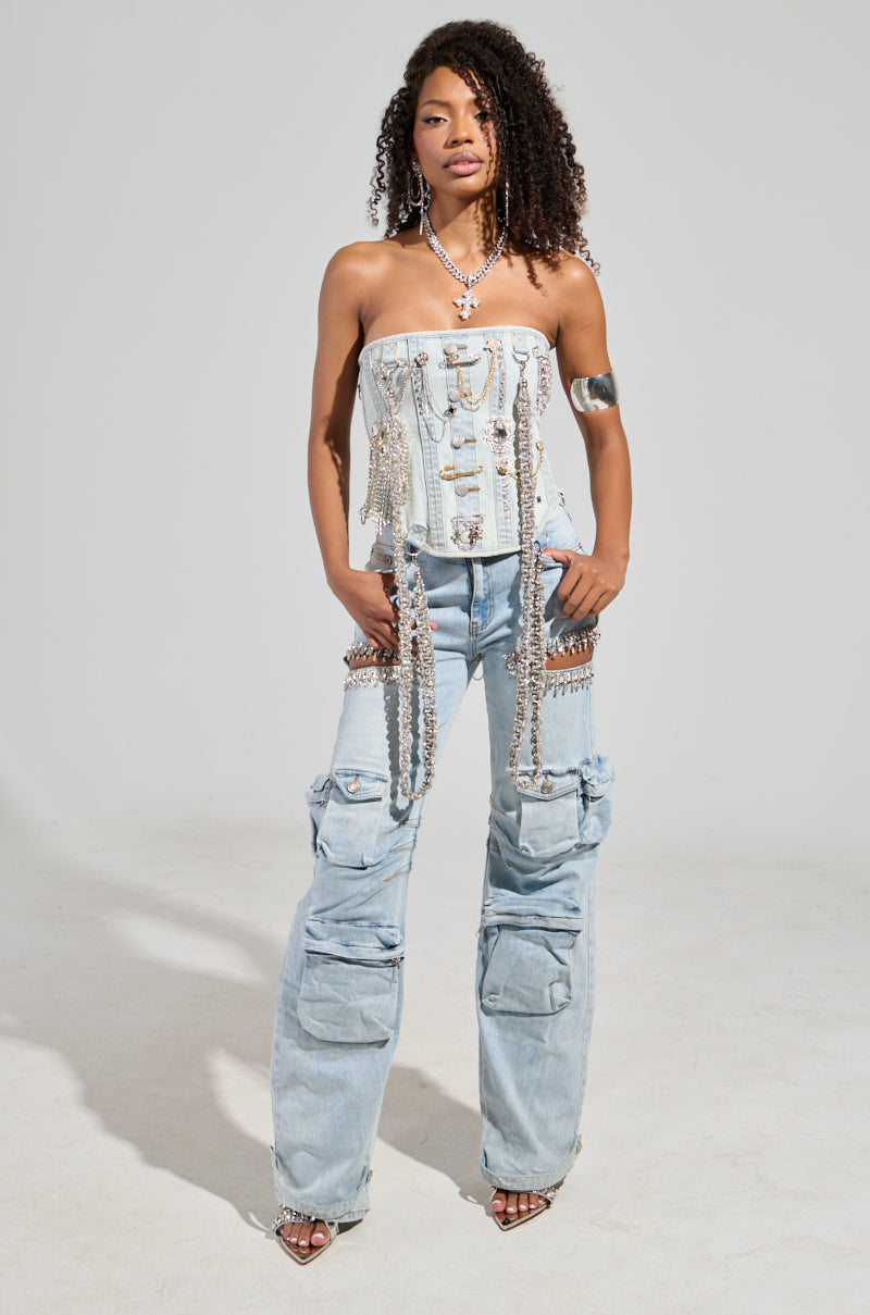 A woman confidently stands against a neutral background, wearing the CALLING THE SHOTS CARGO DENIM WITH RHINESTONES in light blue, featuring a high-rise waist, paired with a strapless top embellished with chains and jewels. Her curly hair complements her necklaces, bracelets, and rhinestone-studded heels.