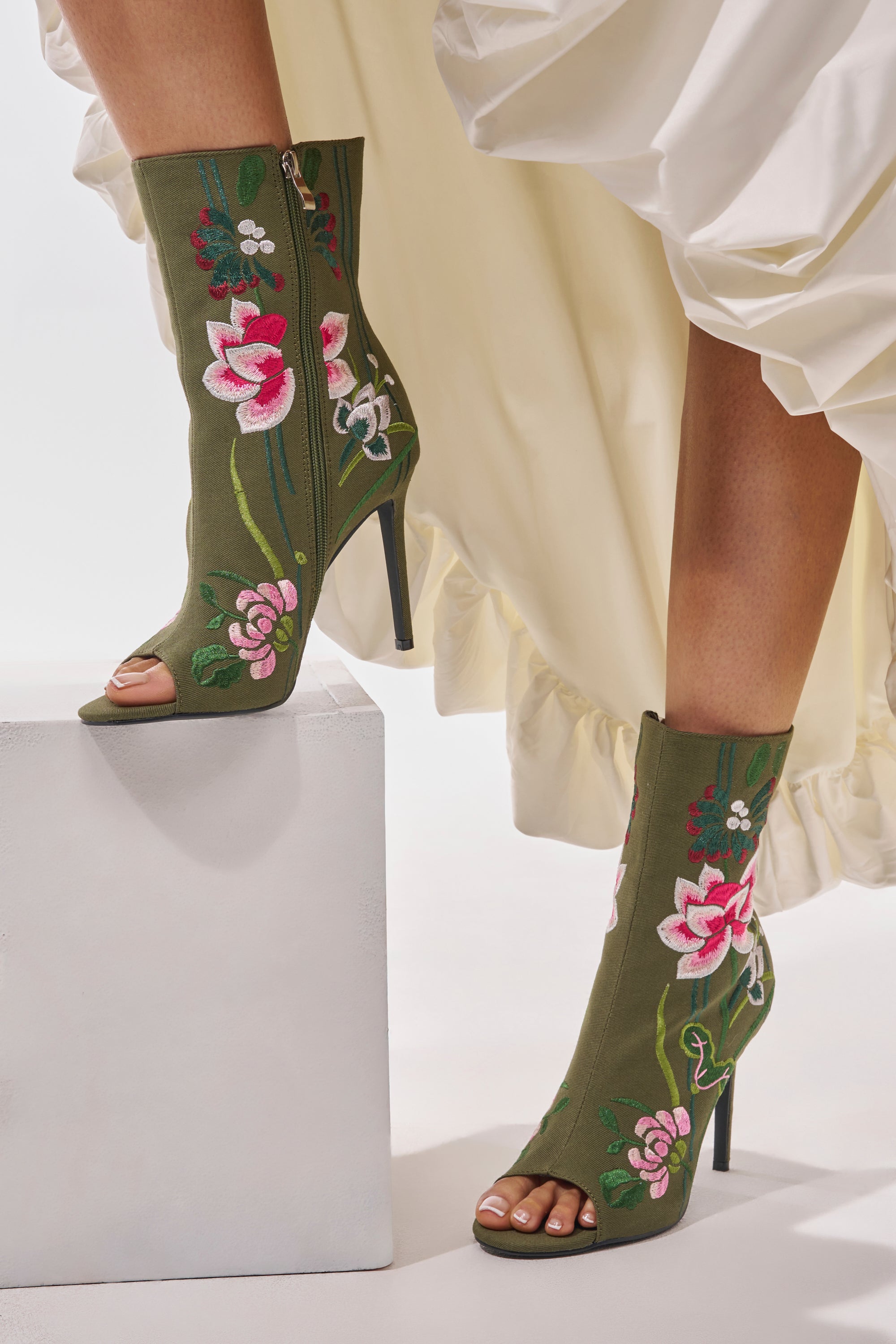 A person wearing the AZALEA WANG DENEVUE OLIVE OPEN TOE BOOTIE, featuring pink and white floral embroidery, stands by a white block with part of a cream ruffled dress showing.