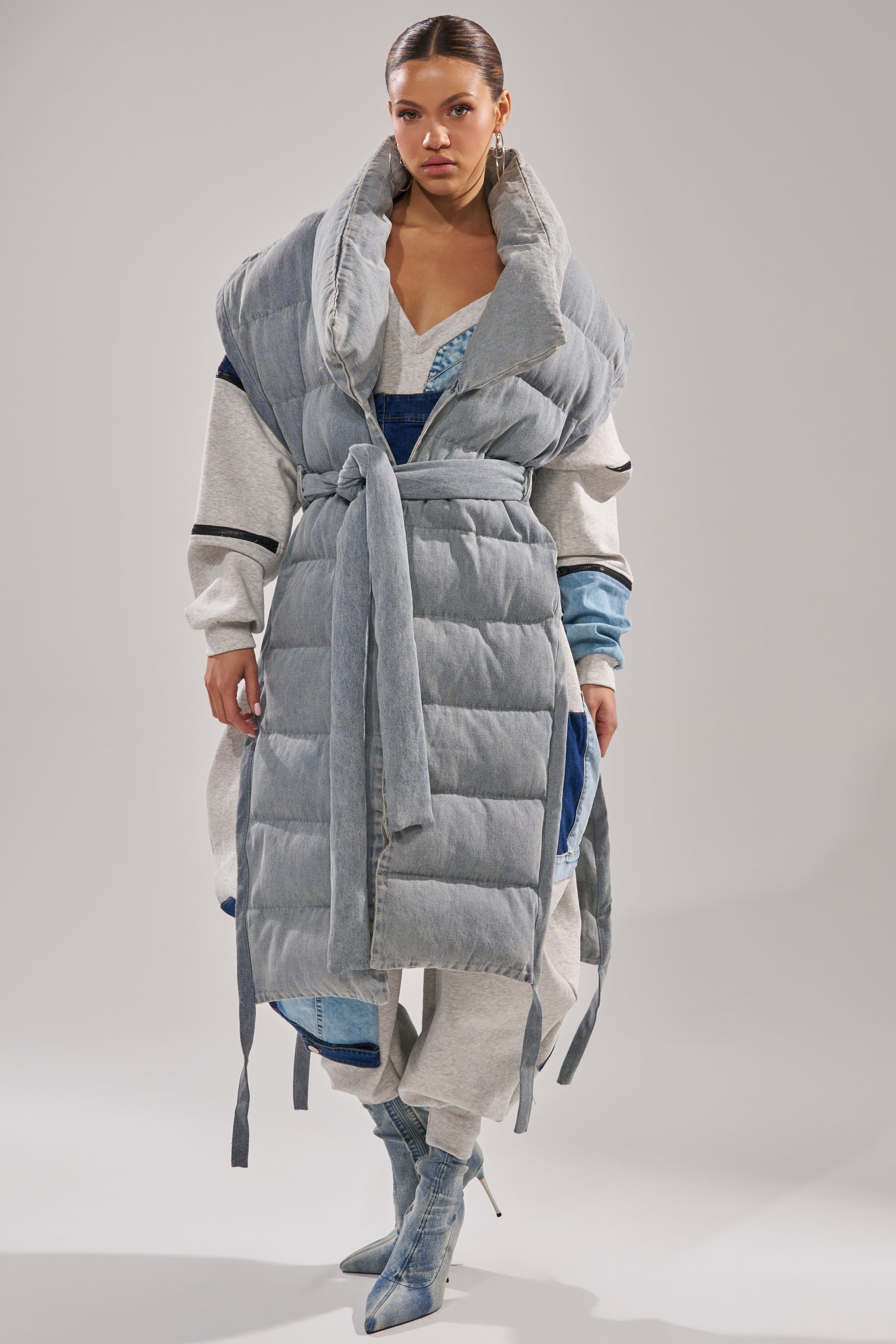 A woman models the PILOT JONES LONG LINE PUFFER VEST IN DENIM, belted at the waist over a gray tracksuit and blue boots, against a plain light background.