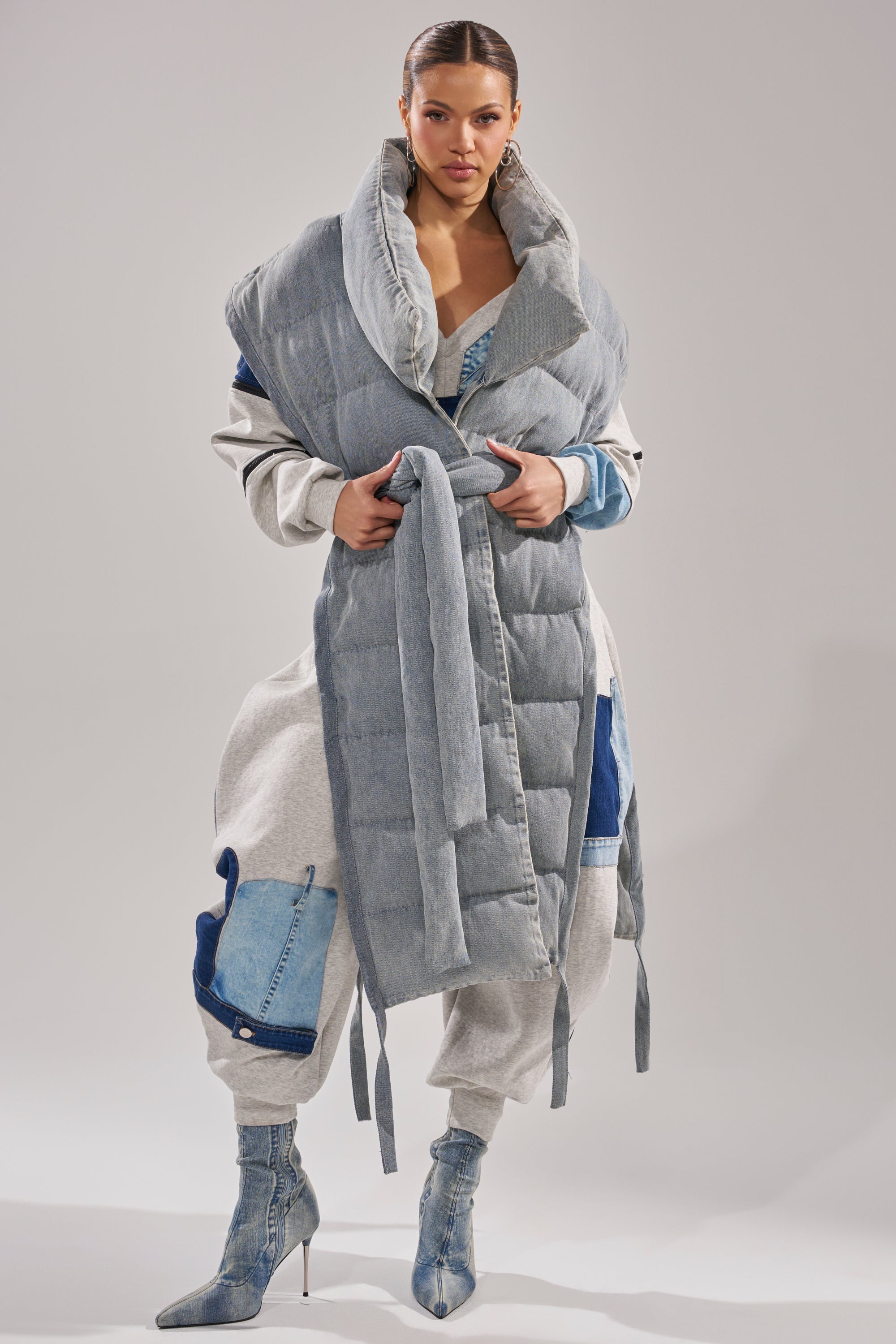 A woman exudes confidence in a grey wrap coat with denim details, layered over the PILOT JONES LONG LINE PUFFER VEST IN DENIM, and styled with matching belted waist and pointed denim ankle boots against a minimalist background.