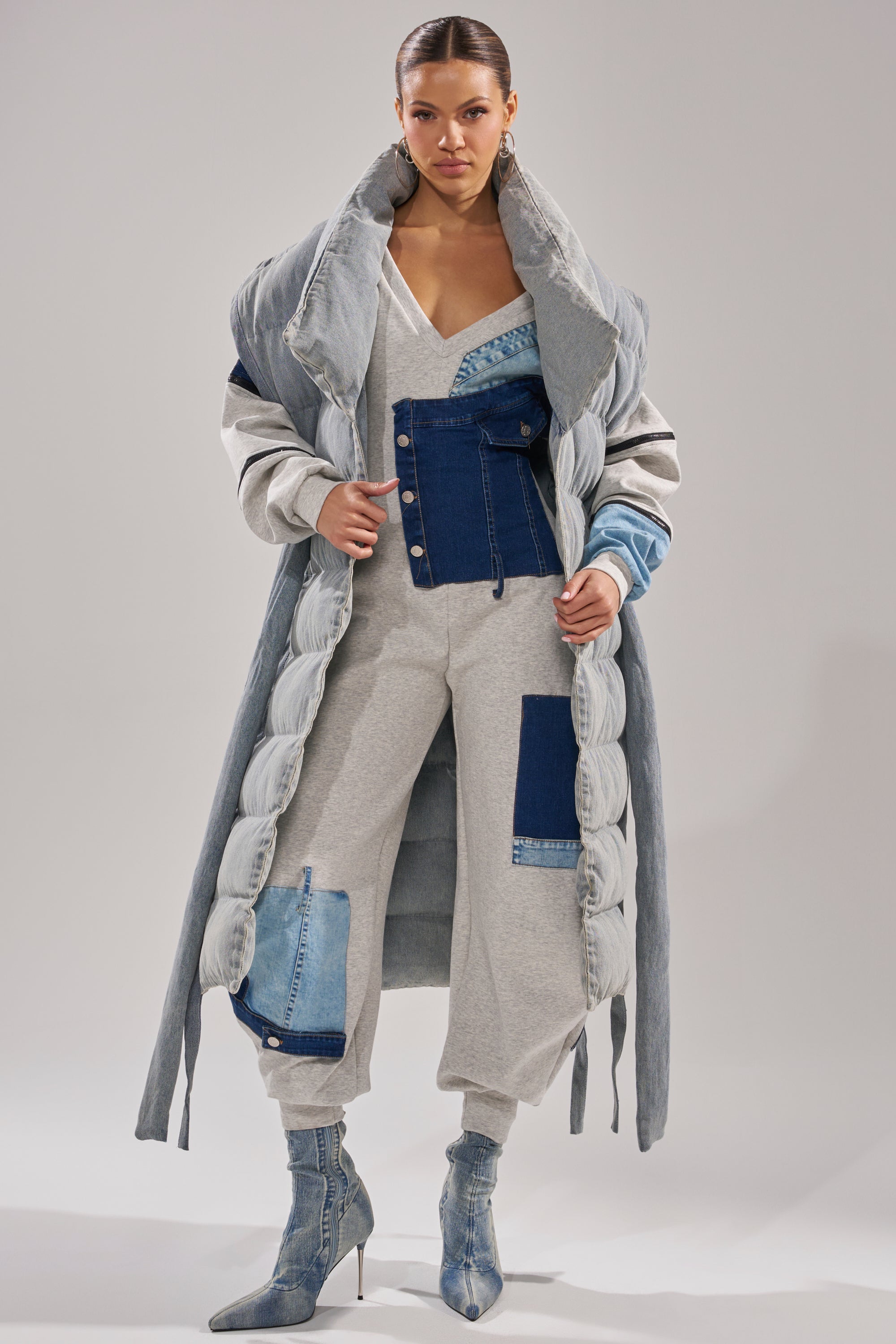 A woman showcases the PILOT JONES LONG LINE PUFFER VEST IN DENIM, layered over a matching patchwork jumpsuit and denim heeled boots, standing confidently against a plain light background.