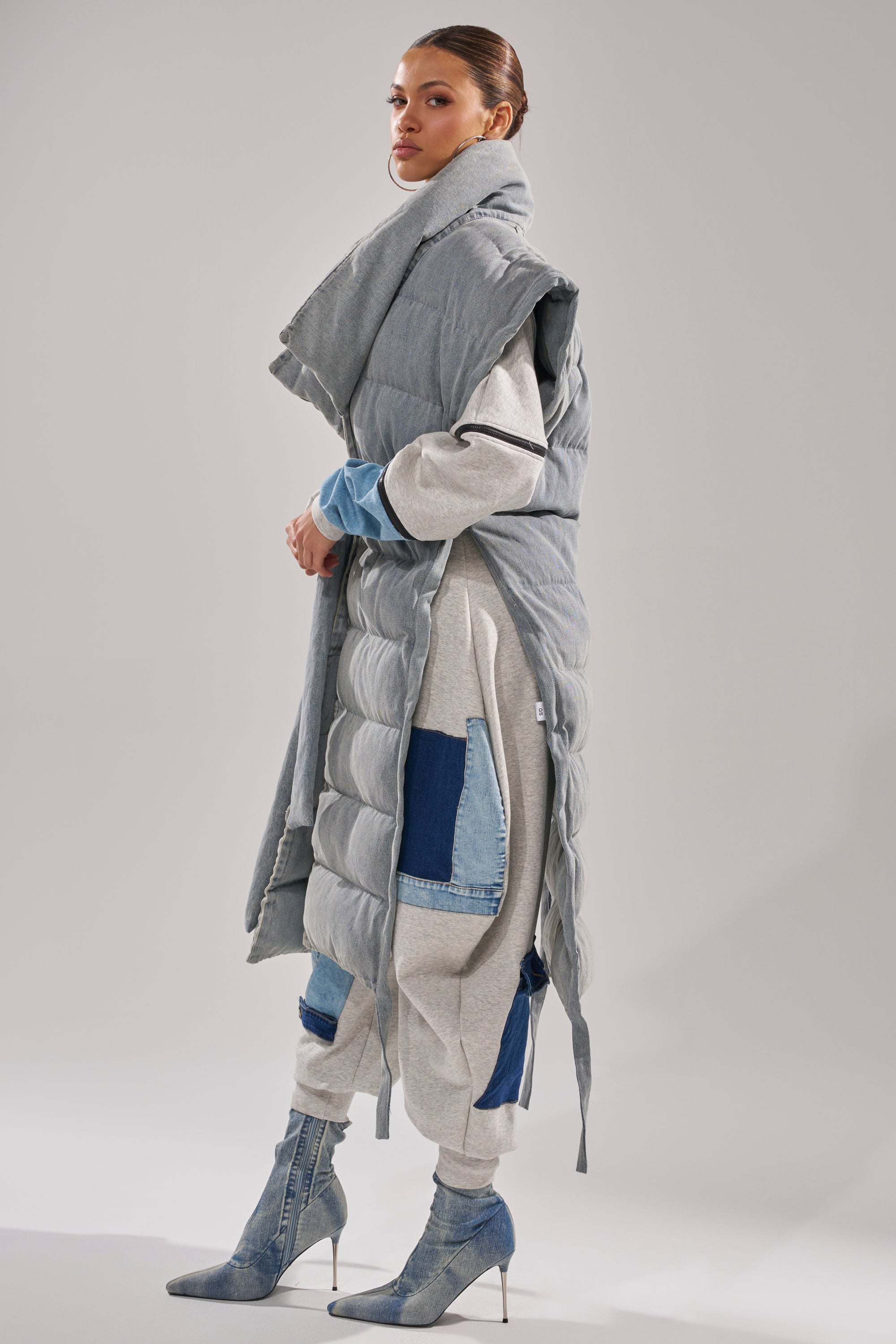 A woman stands in profile wearing the PILOT JONES LONG LINE PUFFER VEST IN DENIM over a light grey tracksuit with blue denim patches, styled with denim heeled boots and slicked-back hair against a plain light grey background.
