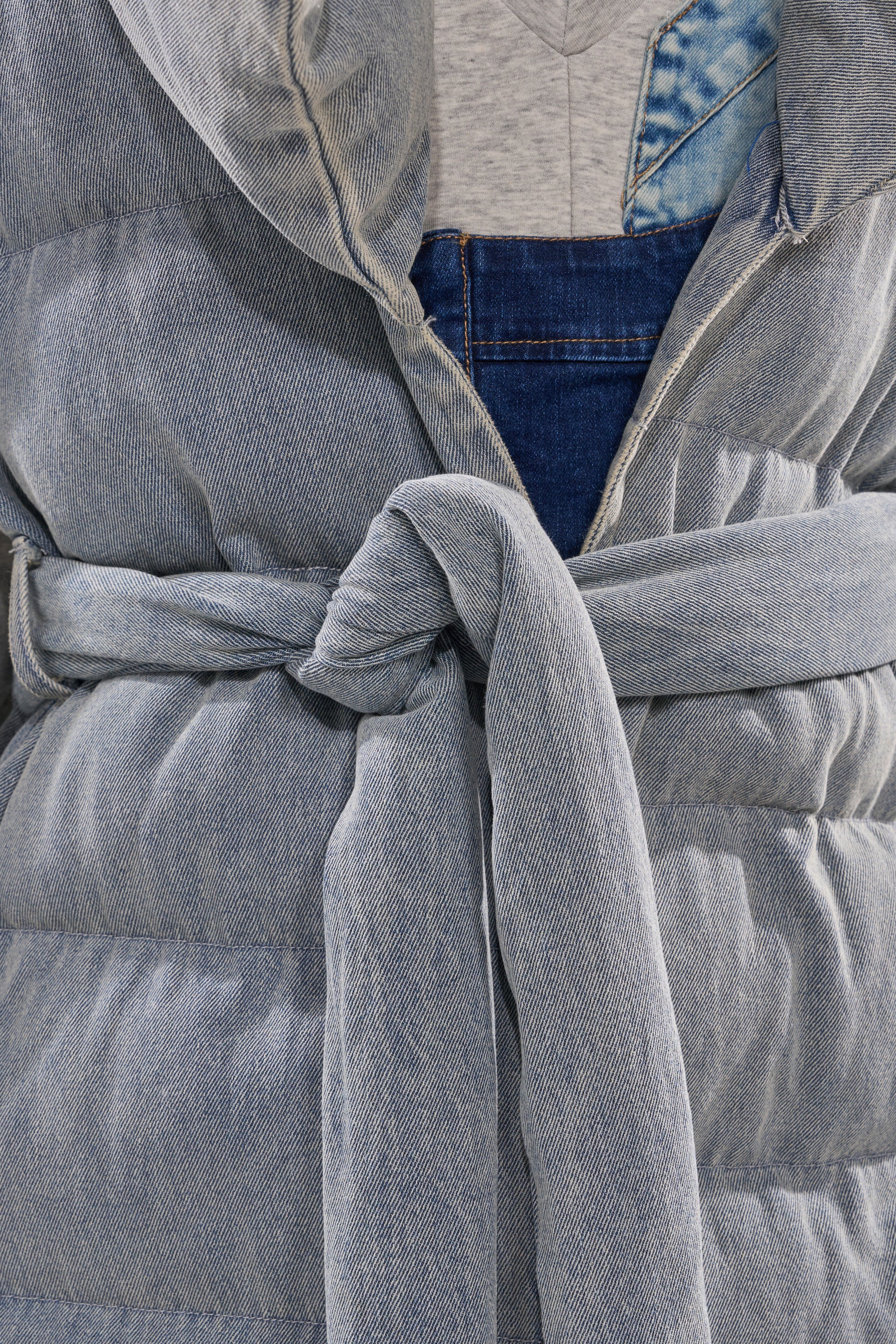 Close-up of a person wearing the PILOT JONES LONG LINE PUFFER VEST IN DENIM over a gray top and blue denim overalls, with the vest’s belt tied in a knot at the waist.