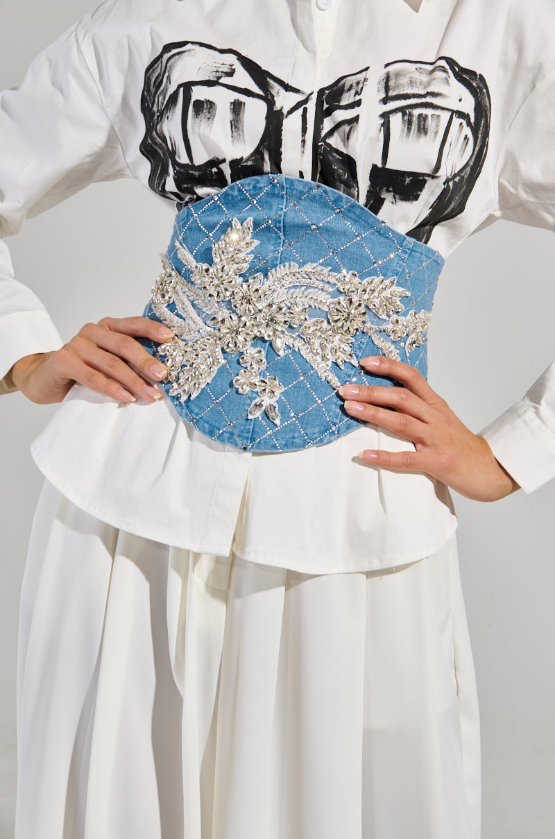 A person wears a white shirt featuring a black brushstroke design on the chest, paired with the TEE EMBELLISHED DENIM BELT, which is wide and blue with intricate rhinestone floral embroidery. The ensemble is completed with a flowing white skirt.