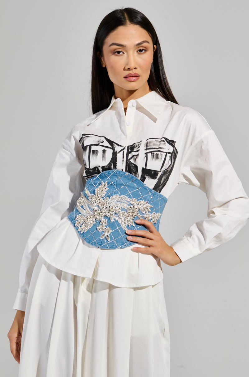 A woman poses confidently in a stylish outfit, featuring a white blouse with a graphic print and the Tee Embellished Denim Belt. Her long dark hair is straight and loose, set against a plain gray background.