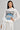 A woman poses confidently in a stylish outfit, featuring a white blouse with a graphic print and the Tee Embellished Denim Belt. Her long dark hair is straight and loose, set against a plain gray background.