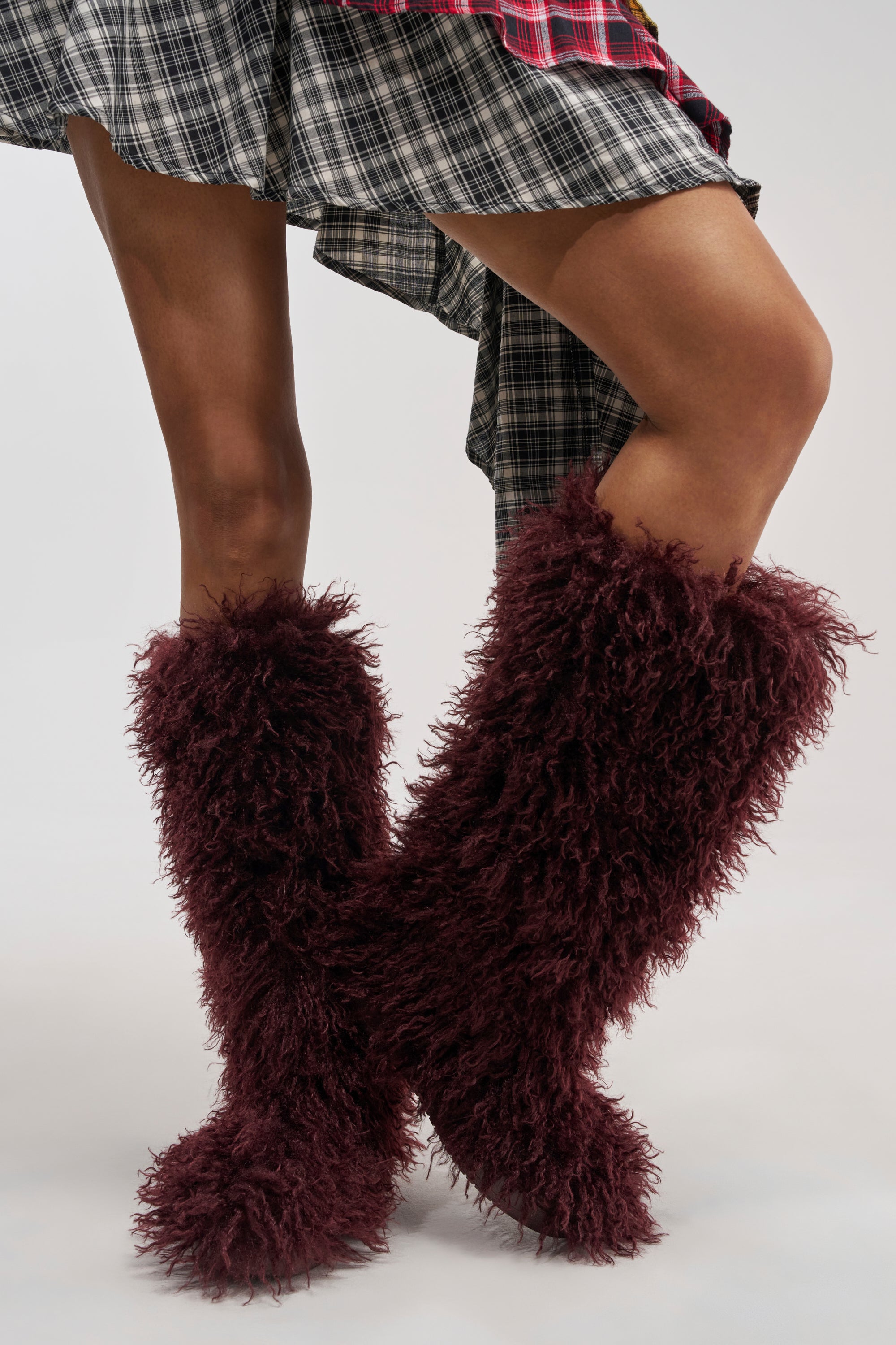 A person wearing a plaid skirt and striking the AZALEA WANG DENTON BURGUNDY KNEE HIGH FLAT FUR BOOTS stands with legs crossed against a plain background.