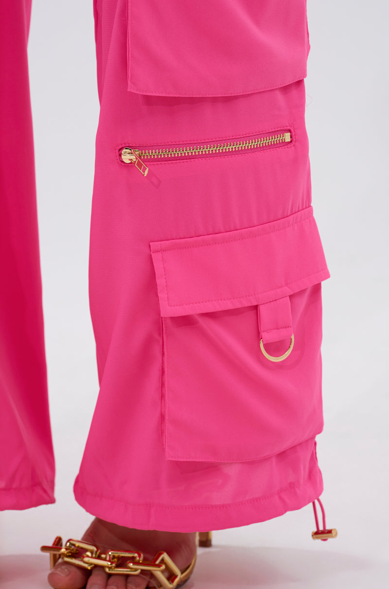 A close-up of someone wearing the UP TO NO GOOD CARGO PANT IN PINK, featuring high-rise, lightweight, slightly shiny fabric with zippered and flap pockets with a gold ring, paired with gold chain sandals. 9
