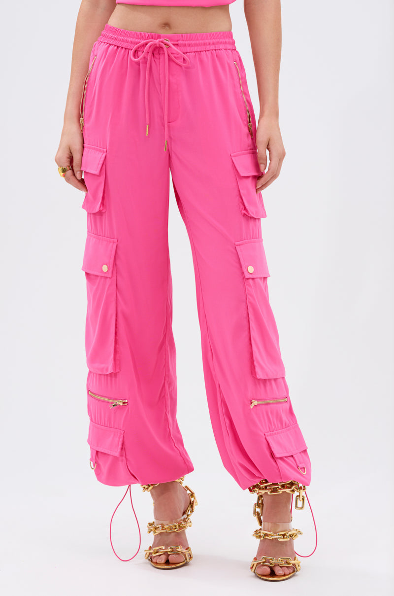 Wearing the UP TO NO GOOD CARGO PANT IN PINK with multiple pockets and zippers, styled with gold chain-embellished heeled sandals. The photo captures the look from waist to feet against a plain background. 1