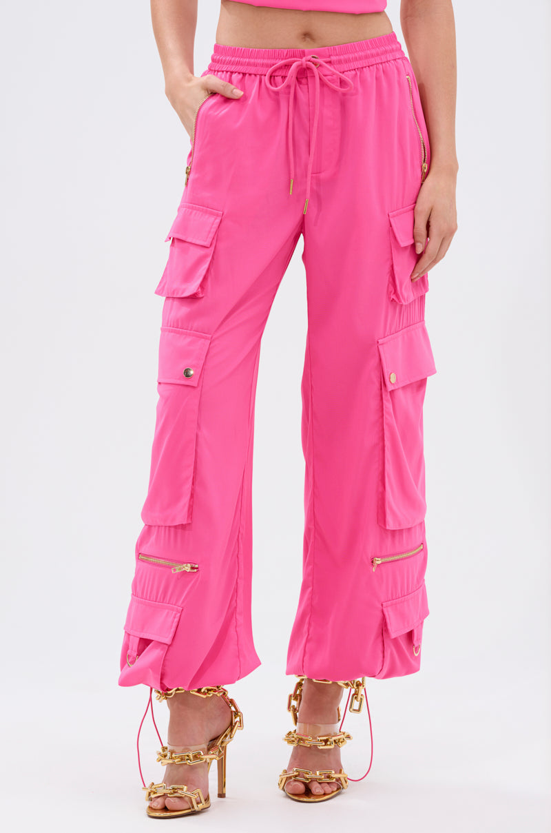 The lower torso and legs of a person in UP TO NO GOOD CARGO PANT IN PINK, featuring multiple pockets and zippers, paired with gold chain-strap high-heeled sandals, are shown against a plain background. 2