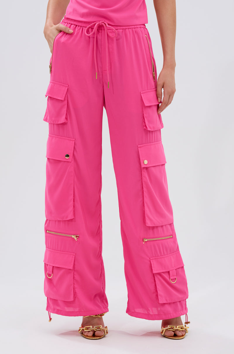 A person models the UP TO NO GOOD CARGO PANT IN PINK—high-rise pants with multiple pockets, gold zippers, and drawstrings—paired with gold chain-strap sandals. The outfit is shown from waist to feet against a plain background. 3