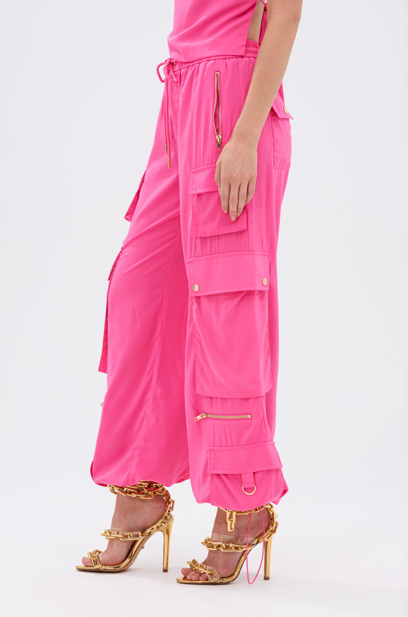Someone wearing the UP TO NO GOOD CARGO PANT IN PINK, a high-rise pink cargo pant with multiple pockets and gold zippers, paired with gold chain-detail high-heeled sandals against a plain white background. 4