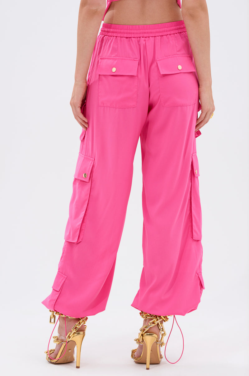 Someone is shown from the back against a plain light background, wearing the UP TO NO GOOD CARGO PANT IN PINK—high-rise cargo pants with multiple pockets and gold button details—paired with strappy gold high heels. 5