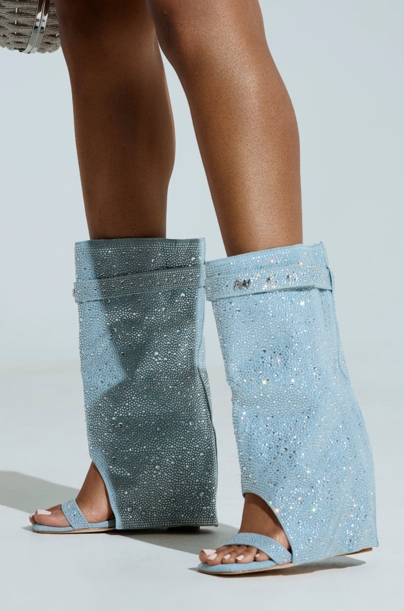 A person wears the AZALEA WANG DESARAY RHINESTONE OPEN TOE WEDGE BOOTIE, featuring shimmering light blue rhinestones and a high, structured design extending up the calves. The boots have a perforated texture and sparkling details against a plain background.