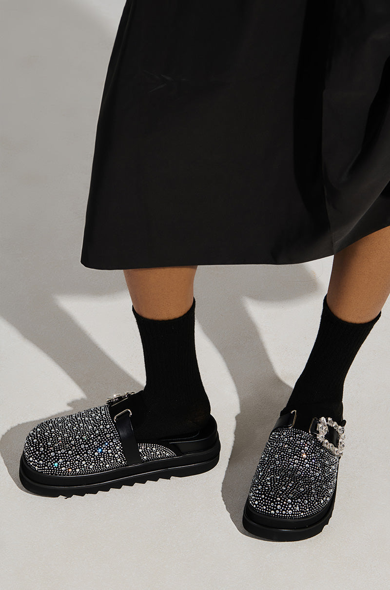 A person wearing black socks and the AZALEA WANG DESDEMONA BLACK FLAT shoes, featuring a strap and rhinestone embellishments, stands on a light-colored floor. The faux suede of the flatform sole adds texture, while the bottom of a black skirt is partially visible above the shoes.