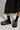 A person wearing black socks and the AZALEA WANG DESDEMONA BLACK FLAT shoes, featuring a strap and rhinestone embellishments, stands on a light-colored floor. The faux suede of the flatform sole adds texture, while the bottom of a black skirt is partially visible above the shoes.