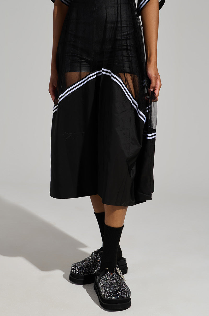 A person is wearing a black mesh skirt with white stripes, black socks, and the AZALEA WANG DESDEMONA BLACK FLAT shoes that feature sparkling studs and a rhinestone embellished heel. The background is plain and neutral.