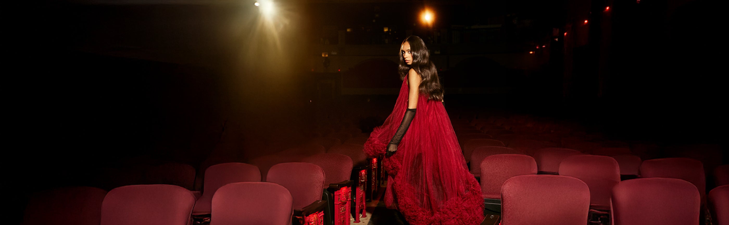 Woman in a red dress standing on a theater stage with red seats and spotlights.