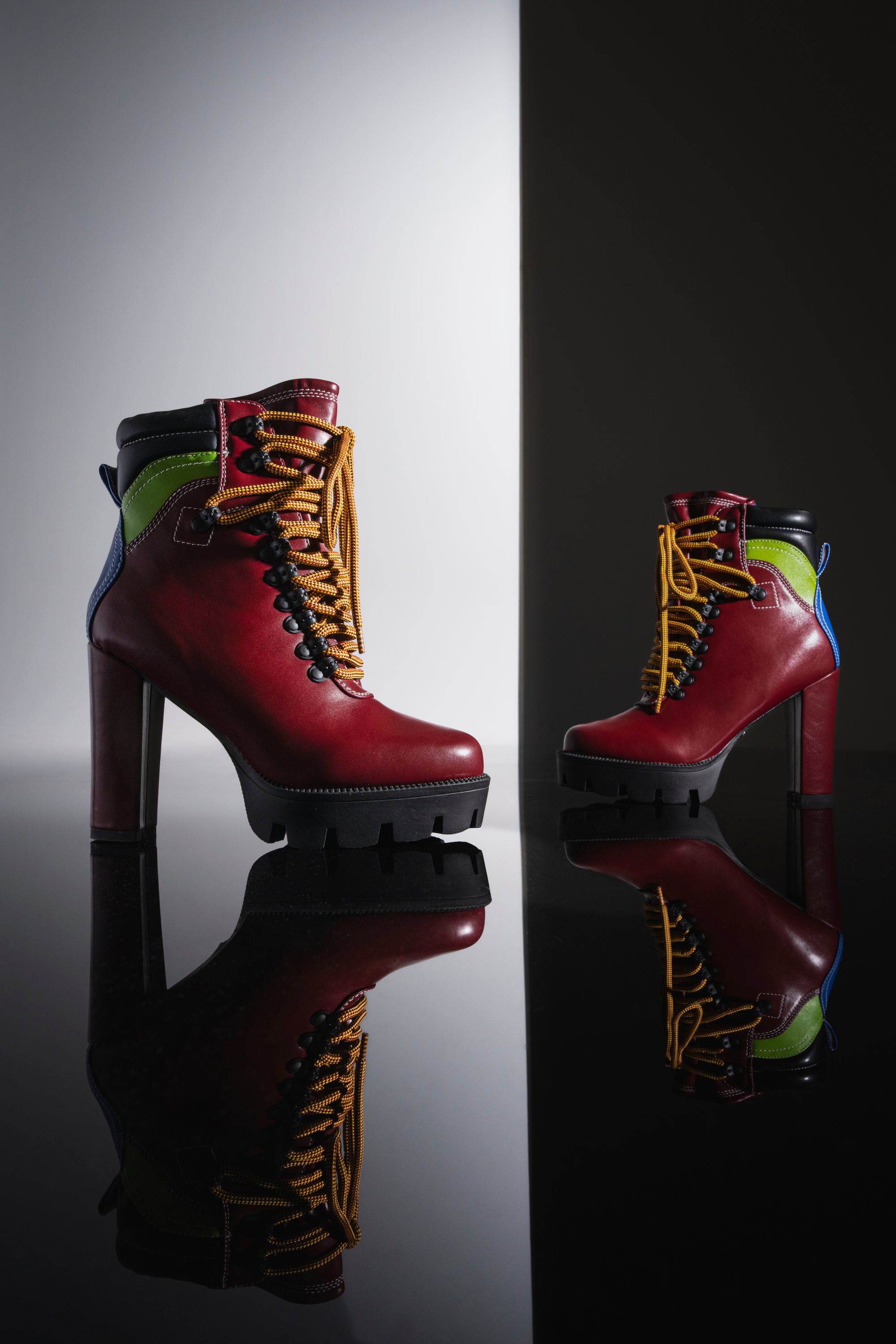 The AZALEA WANG DEUTCH MULTI SPORTY BLOCK BOOTIE is a pair of high-heeled, lace-up streetwear boots in vibrant red leather, featuring striking yellow laces and bold green and blue accents. The boots are showcased with their reflection on a glossy black surface against a split black and white background.