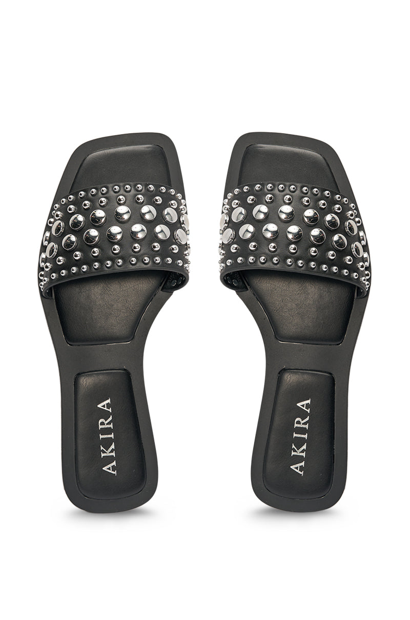 The AKIRA DEVORA BLACK HARDWARE EMBELLISHED FLAT SLIP ON SANDAL features black wide straps with assorted silver studs, square open toes, black faux leather footbeds, and "AKIRA" in white lettering. 3